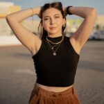 A stylish young woman poses confidently outdoors at sunset, embracing a casual summer vibe.